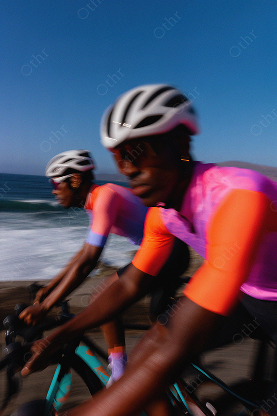 Two Cyclists Riding Along Coastal Road With Motion Blur and Vibrant Jerseys