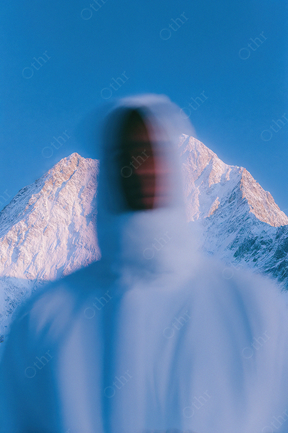 Blurred Figure in Hood Standing Before Snowy Mountain Peak at Blue Hour