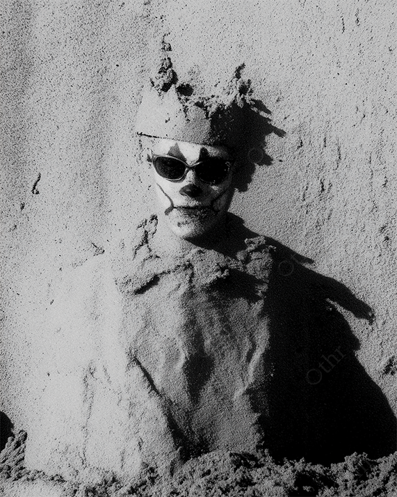 Clown Face with Sunglasses and Sand Crown Buried in Wall of Dry Sand – Black and White