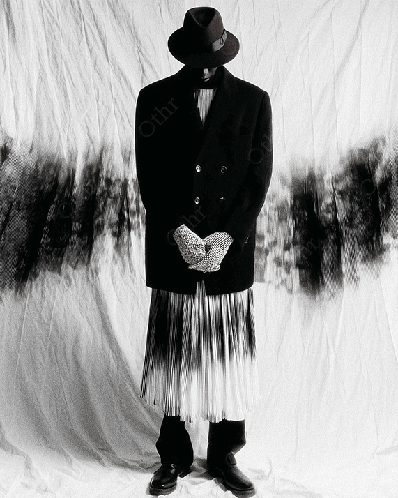 Model in Fedora and Pleated Skirt Posing Against Draped Backdrop with Ink-Like Patterns – Monochrome