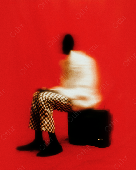 Blurred Figure in White Jacket and Chequered Trousers Sitting on Television Against Red Background