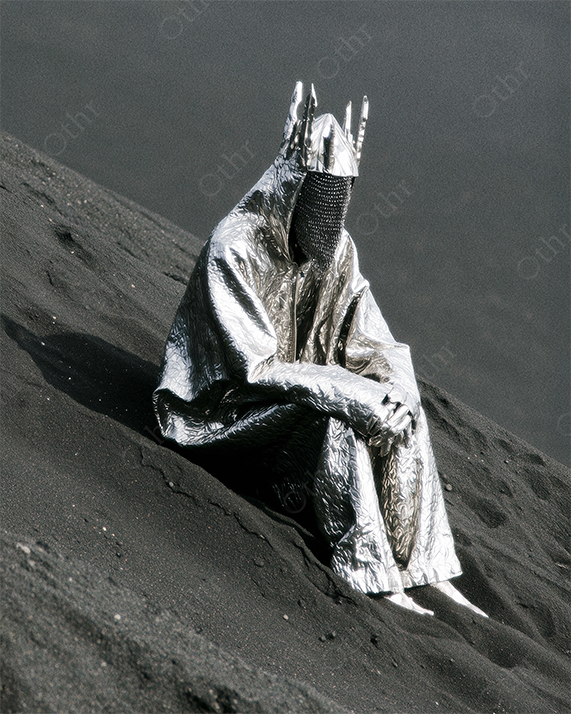 Figure in Metallic Cloak and Crowned Helmet Sitting on Dark Sand Dune