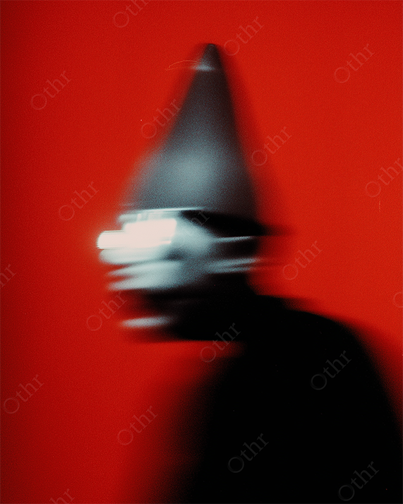Blurry figure wearing a sharp conical hat and reflective eyewear on a red background.