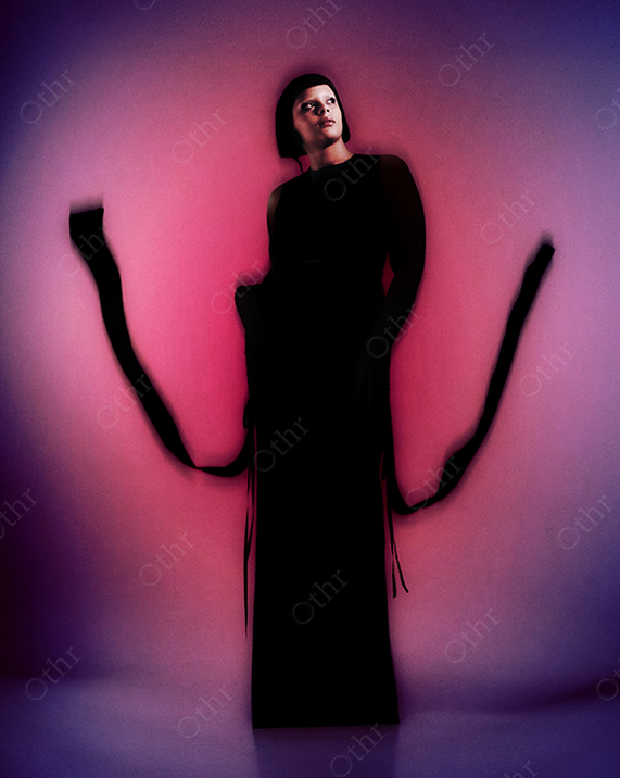 Person Standing in Long Black Dress With Flowing Ribbons Against Pink and Purple Gradient Light