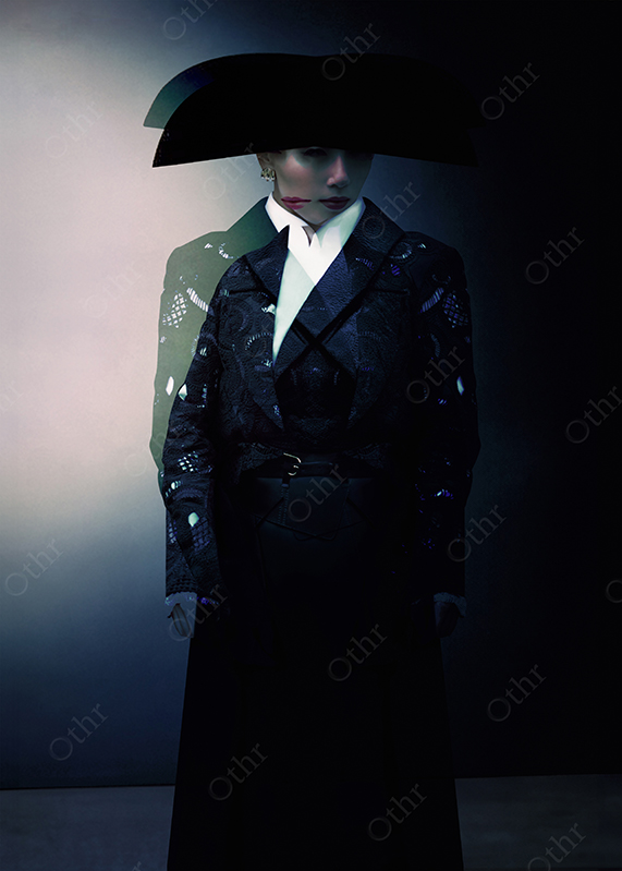 Studio Portrait of Woman Wearing Wide-Brim Hat and Patterned Coat Standing in Low Light Against Neutral Background