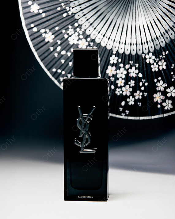 Black Perfume Bottle With Metallic Logo Positioned in Front of Decorative Hand Fan Against Dark Background