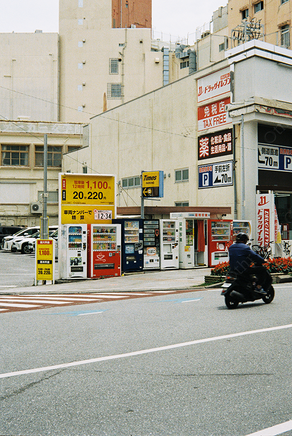 Urban Street Corner With Vending Machines, Parking Signage, and Passing Scooter in Daylight