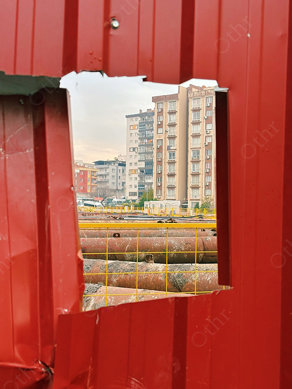 Apartment Buildings Seen Through Torn Red Metal Fence at Construction Site
