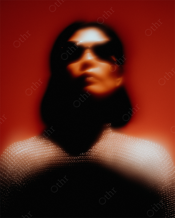 Blurred stylised portrait of woman wearing sunglasses and chainmail in red light.