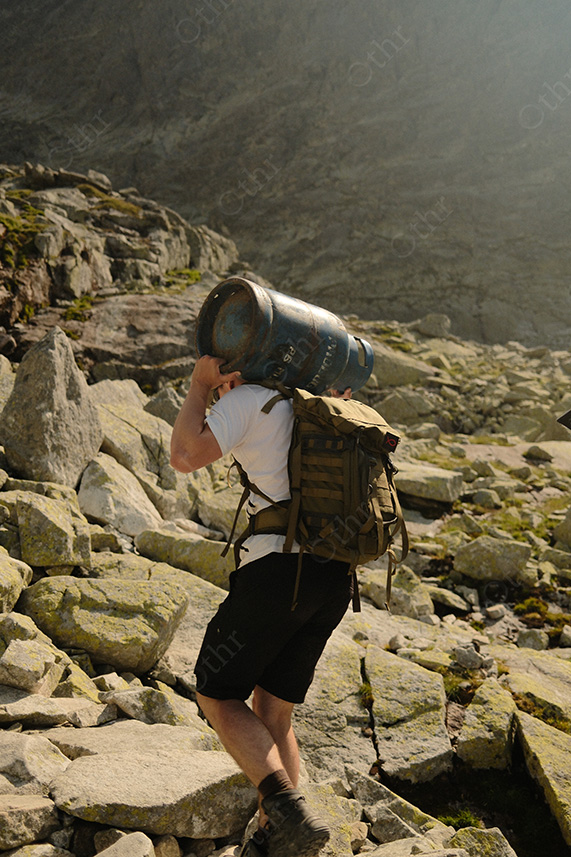 Hiker Carrying Metal Keg on Rocky Mountain Trail in Harsh Sunlight