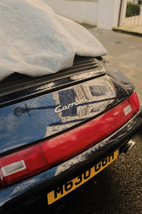 Close-Up of Car Rear With “Carrera” Badge and Raindrops Beneath Grey Car Cover