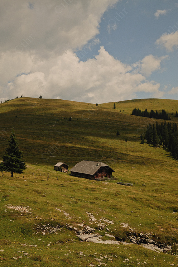 Isolated Mountain Cabin on Sloping Meadow Beneath Dramatic Clouds and Blue Sky