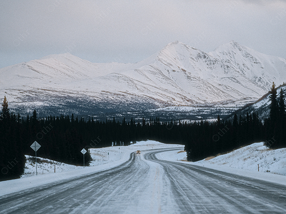 Snow Covered Road Leading Towards Mountain Range Under Overcast Sky