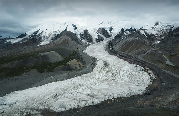 Glacier Flowing Through Mountain Valley Beneath Snow Covered Peaks Under Cloudy Sky