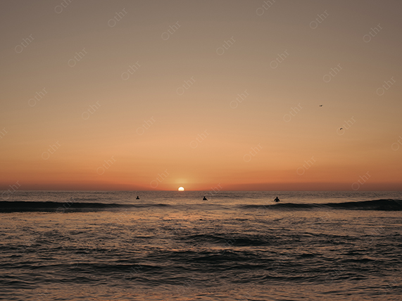 Sun Setting on Ocean Horizon With Surfers Silhouetted Against Orange Sky