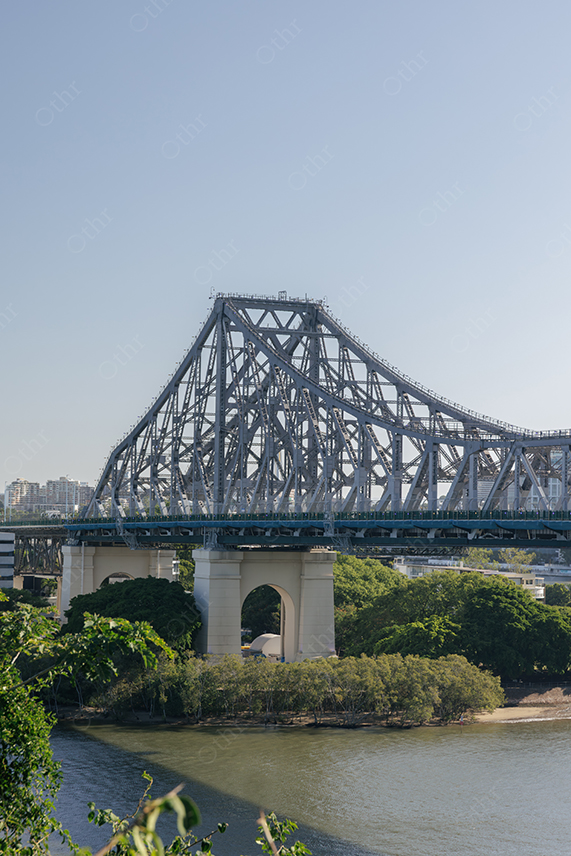 Large Steel Bridge Over River With Trees and Cityscape Under Clear Blue Sky